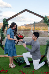 Man proposes to woman with ring and flowers under a neon 'Will You Marry Me' sign