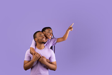 Obraz premium Portrait Of Excited Black Young Couple Hugging, Looking And Pointing Finger Up And Aside At Free Copy Space On Purple Violet Background, Lady Piggybacking Guy, Demonstrating Place For Advert, Banner