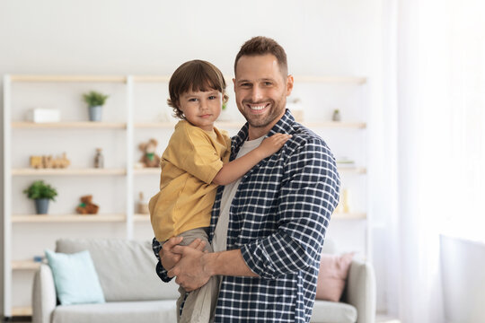 Family portrait. Young positive single father holding his little son on hands and smiling to camera, posing together at living room interior