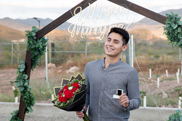 Man holding a bouquet of red roses and an engagement ring box, proposing marriage outdoors