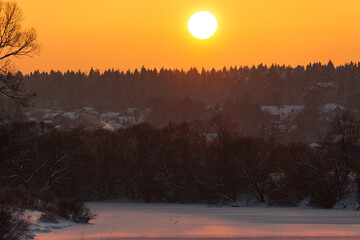 Fototapeta premium Vibrant winter sun bathes a serene frozen river and snow-dusted trees. Distant village homes peek through the atmospheric golden evening glow. Idyllic landscape