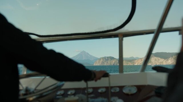 Close-up of skipper's hands at ship's helm and control panel in yacht's wheelhouse with view out window of mountains and volcano on bay coast. Blurred foreground