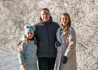 Fototapeta premium Smiling parents with their young daughter standing in sunny winter forest. Ice glazed trees around, family bonding, joyful mood, warmth and affection contrasted with frosty outdoor scenery.