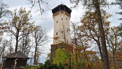 A partially plastered stone fortress tower with a balcony and windows stands among oak trees with yellowed foliage. There is a gazebo and lampposts nearby. Cloudy autumn weather