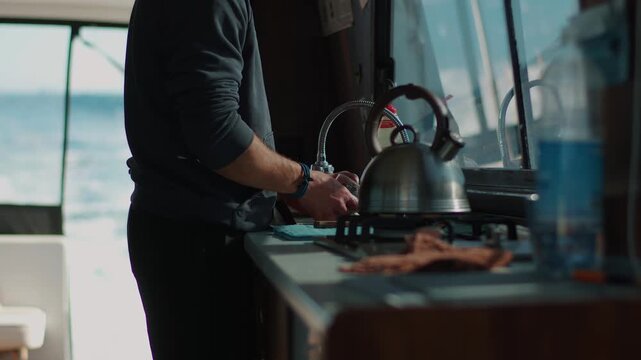 Brutal man with sleeves of black jumper rolled up washes dishes in yacht's galley next to stove with kettle on it. Close-up of his hands. Blurred background of sea bay is behind him