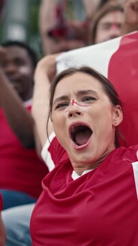 Caucasian female football fan with red and white face paint clasping hands looking up emotionally during match. Woman in red jersey watching game anxiously in crowded stadium. Outdoors. Vertical.
