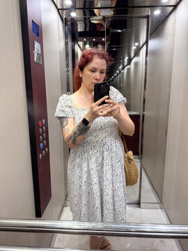 Woman in white dress taking selfie in elevator mirror with round straw bag. Fashion, summer style, and lifestyle identity