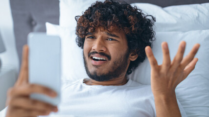 A young man lies on his bed and looks at his smartphone with a puzzled expression. He raises his hands in confusion. The room is well-lit during the day.