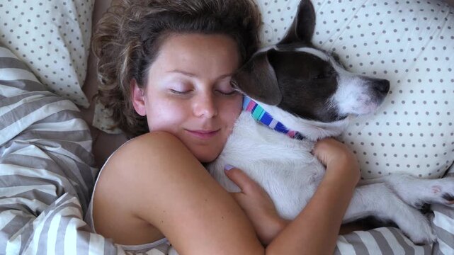 Adorable top view of a young smiling woman cuddling her cute dog while sleeping together in a cozy bed, representing a strong bond between human and pet, love, and companionship