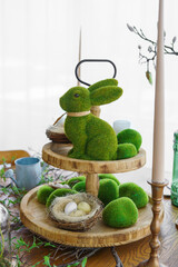 Green moss Easter Bunny and decorative Easter eggs in nest on on a two tiered wooden plate in home background. Home decor for spring holidays, festive table setting