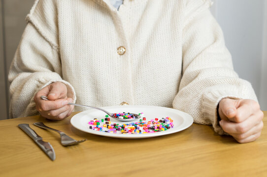 A person sitting at a table holding a spoon filled with colorful plastic beads over a plate, representing the human intake of microplastics.