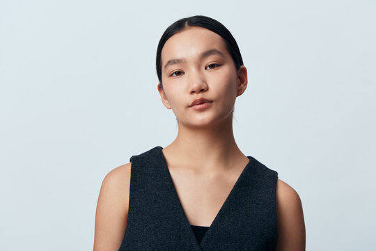 Portrait of young woman with neutral expression wearing dark sleeveless top against plain light background, looking directly at camera in studio setting.