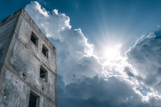 Dramatic sun rays breaking through thick clouds beside an unfinished concrete building under a bright blue sky on a partially cloudy day