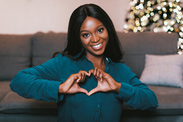 Obraz premium A woman with dark hair smiles at the camera while making a heart shape with her hands. She is sitting on a couch in a living room with a Christmas tree decorated with lights behind her.