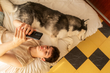 A 17-year-old teenage boy lies in bed with his husky dog ​​and looks at his phone, a concept of a leisurely teenage morning routine