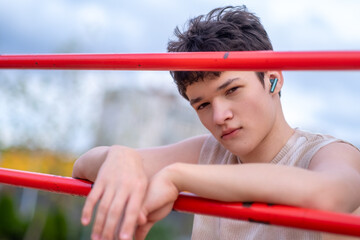 A young person with a serious expression stands outdoors near a sports playground, listening to...