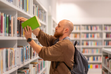 A bearded man selects books in a public library. He picks up a textbook he's chosen. Research and education concept