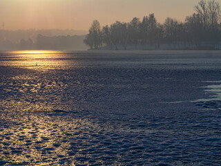 Frozen lake in winter at sunset, golden light reflecting on icy surface, misty background and bare trees creating a calm, serene and atmospheric winter landscape with copy space