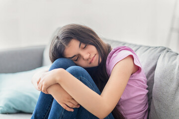 Obraz premium Unhappy Indian teen girl sitting on sofa with closed eyes, hugging her knees, feeling depressed or desperate at home. Lovely asdolescent having problem or teenage depression