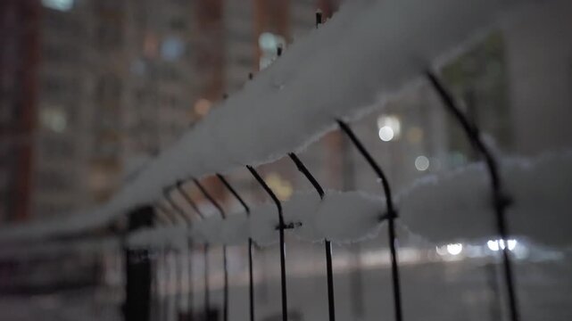 Snow covering a metal fence on a cold winter night with blurred city lights glowing in the background. Atmospheric urban street scene with shallow depth of field and soft bokeh illumination. Quiet cit