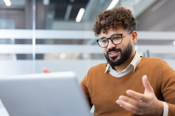 Young bearded man with curly hair and glasses having an online business video call on a laptop, actively communicating and gesturing from a modern office workspace