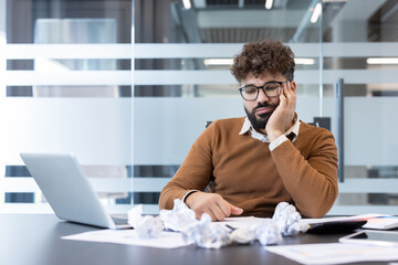 Young man feeling stressed and overwhelmed by work, resting his head on his hand at an office desk with crumpled papers and a laptop, struggling with fatigue and lack of ideas