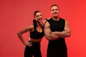 Obraz premium Happy fitness trainer man and woman posing and smiling, both wearing black sportswear, lady putting hand on guy shoulder, standing over red neon background