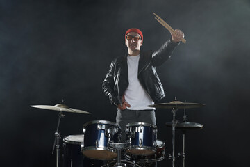 Portrait of smiling man in hat and sunglasses with drumsticks and drum kit in smoke against black...