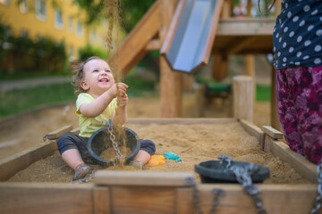 Laughing toddler girl sitting in a sandbox, looking up at sand pouring down on her hands and buckets.