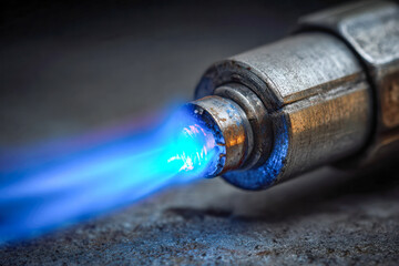 Detailed view of a metallic blowtorch igniting a bright blue flame while resting on a textured surface for industrial or craftsmanship use