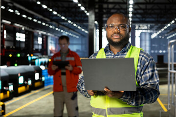 African american supervisor overseeing heavy tools near welding sparks, using laptop with hi vis vest and safety glasses. Manufacturing development and powerful industrial environment.