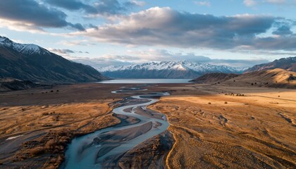 Vast mountain valley landscape with a braided river winding through golden grasslands towards a lake and snow-capped peaks under a dramatic cloudy sky at sunset