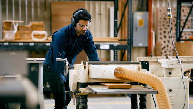 Craftsman using woodworking machinery for planning wood piece in a professional carpentry workshop, wearing safety gear for protection
