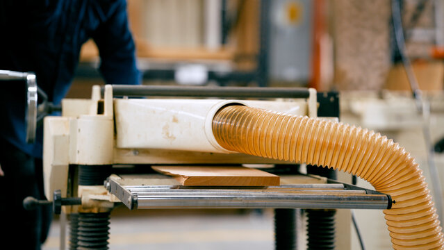 Woodworker planing a timber plank with a thickness planer, collecting sawdust through a dust extraction hose