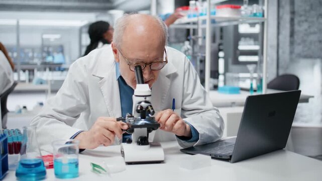 Portrait of cheerful senior lab researcher looking through scientific microscope lens to study cells. Elderly microscopist performing biological sample examination with optical equipment, camera B
