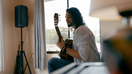Man with long hair sitting by window, concentrating on playing acoustic guitar during a practice session indoors