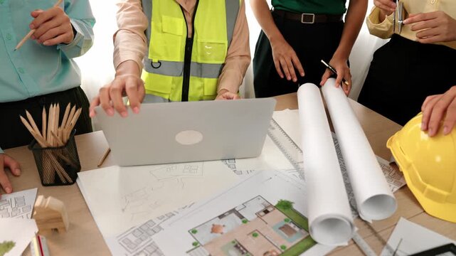 Architect and safety engineer discussion about building blueprint displaying on laptop on table with architectural document and wooden block scatter around at modern office. Closeup. Delineation.