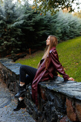 Woman in maroon coat sitting on stone wall in park with greenery