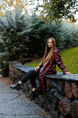 Young woman in maroon coat sitting on stone wall in park setting