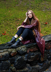 Young woman in maroon coat sitting on stone wall in outdoor park setting