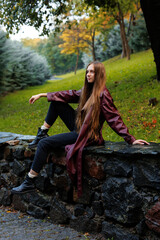 Young woman in maroon coat sitting on stone wall in park setting