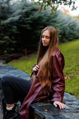 Young woman in long maroon coat sitting on stone wall in park