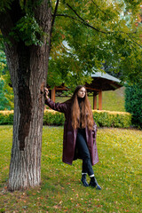 Young woman in long coat leaning against tree in park setting