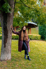 Young woman in long coat leans against tree in park during autumn