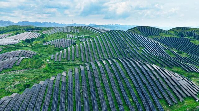 Aerial shot of a massive solar panel farm on green rolling hills generating renewable energy under a cloudy sky.