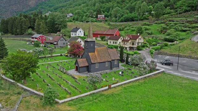 Aerial view of Flam kyrkje and cemetery beside the river in Norway. Drone footage shows historic wooden church surrounded by village homes and green fields.