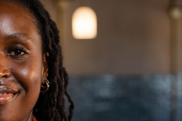 African woman smiling, facing left in cafe, showing gold hoop earring, warm collar, pendant light