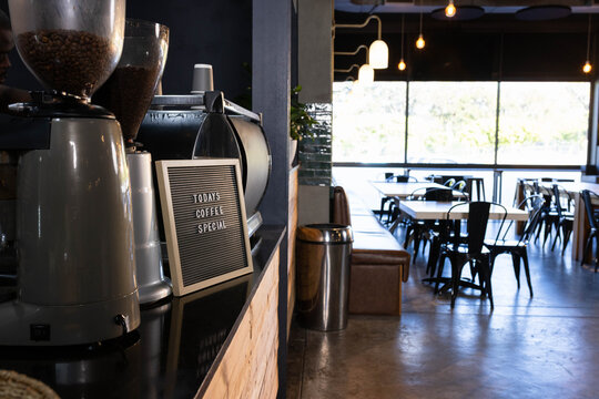 Service counter is holding espresso machine, bean hoppers, letterboard sign by window with greenery