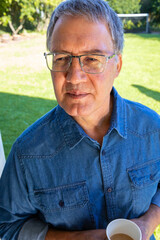Senior man standing on porch wearing denim shirt, glasses holding white cup gazing over sunny lawn