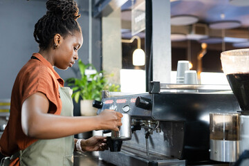 African American woman wearing green apron steaming milk with pitcher at cafe espresso machine